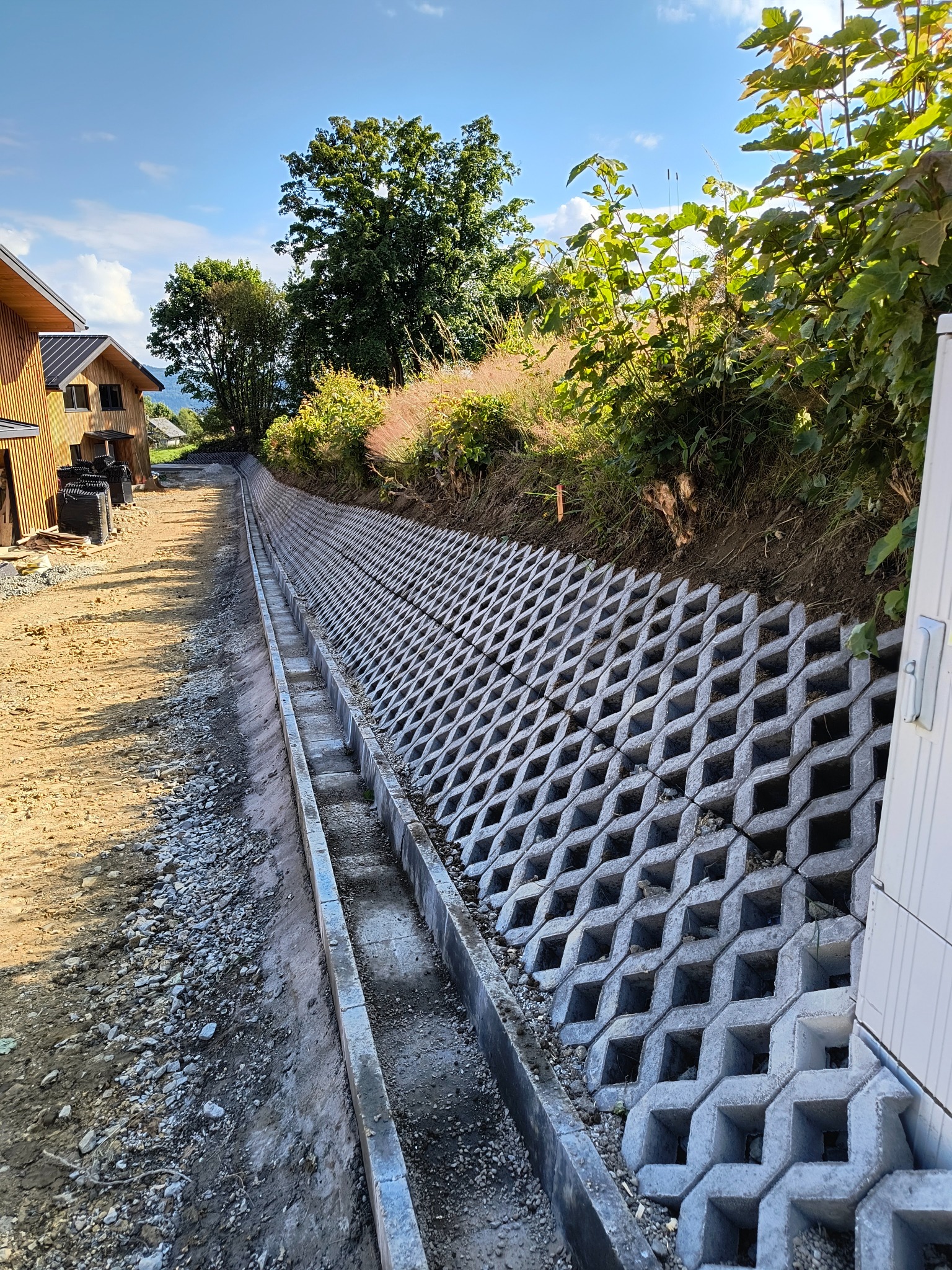 Wzmocnienie skarpy przy drodze za pomocą ażurowych betonowych elementów, widoczny rów odwadniający i fragment drewnianego domu w tle.