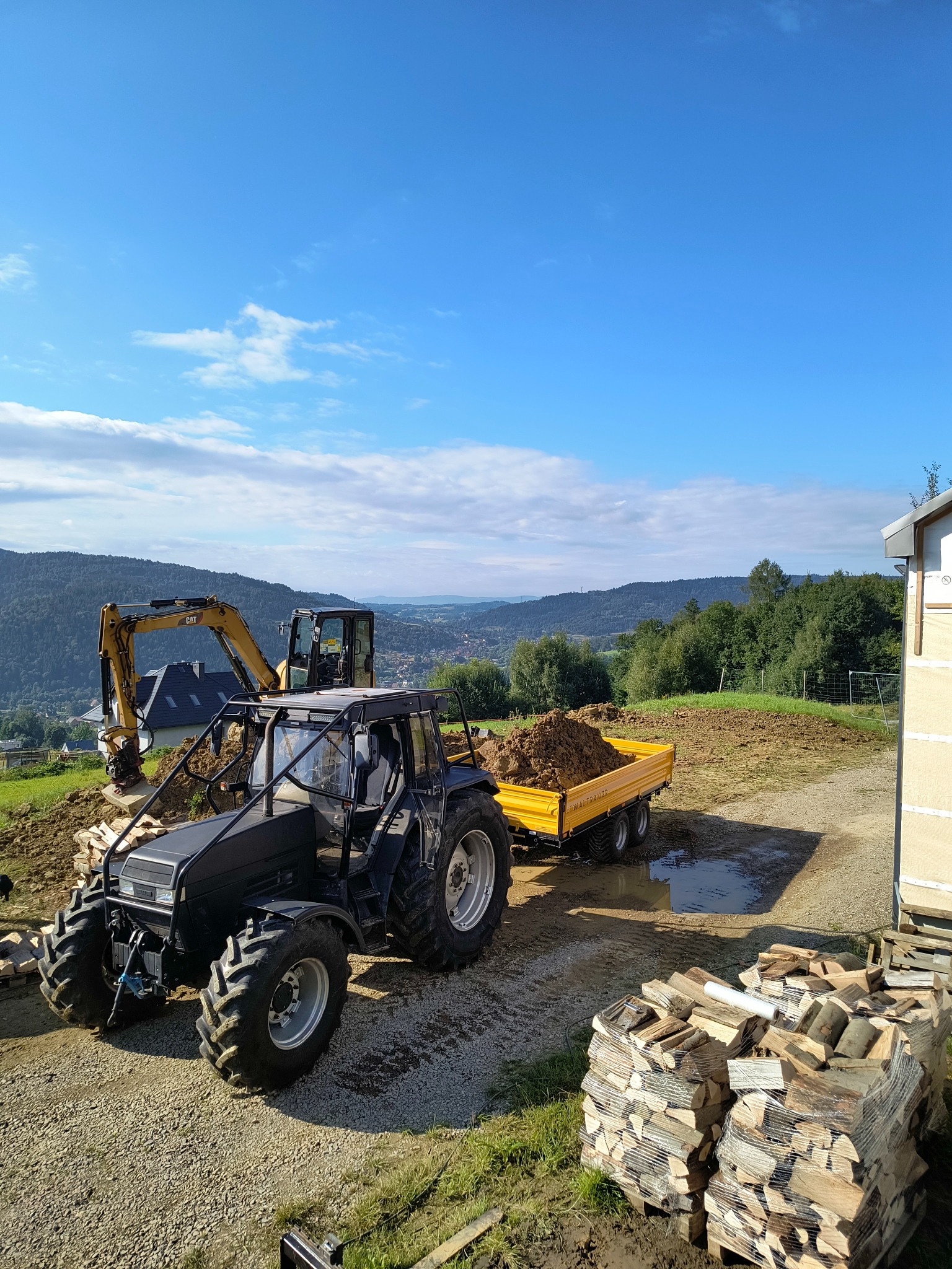 Czarny traktor z przyczepą wypełnioną ziemią, obok koparka CAT, w tle pagórkowaty krajobraz i sterta pociętego drewna na paletach.