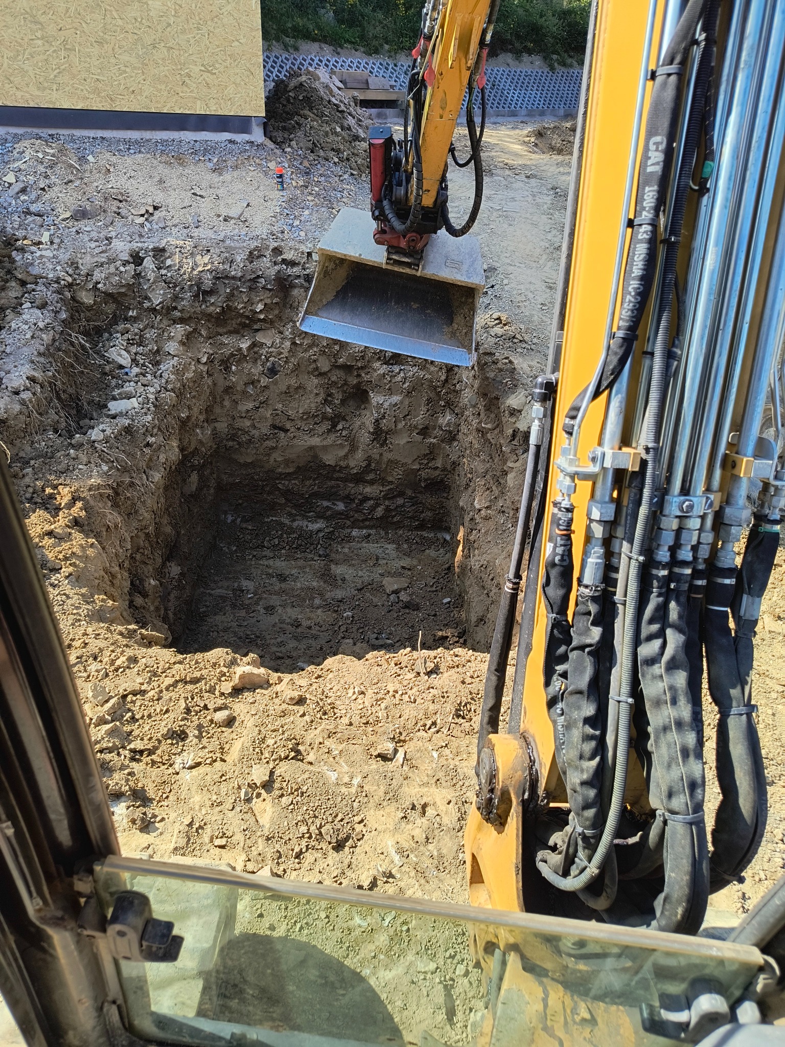 Żółta koparka z łyżką pogłębia wykop w ziemi, widok z kabiny operatora, w tle fundamenty budynku.