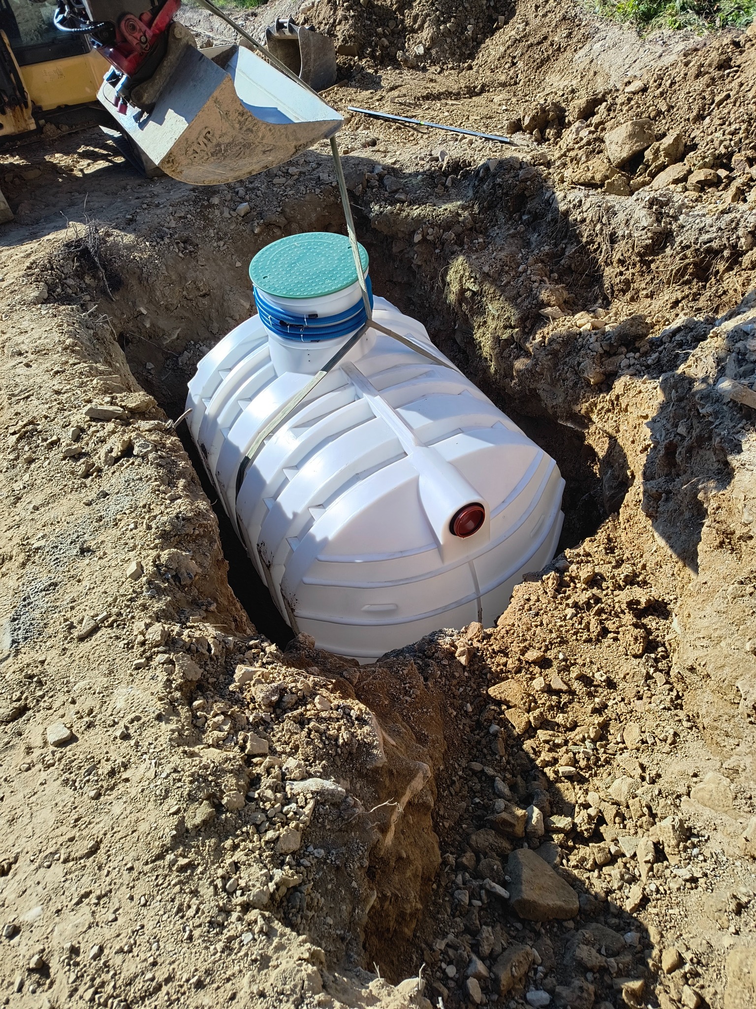 Biały, plastikowy zbiornik z zieloną pokrywą, opuszczany do wykopu za pomocą koparki, widoczne ślady ziemi na zbiorniku i ścianach wykopu.