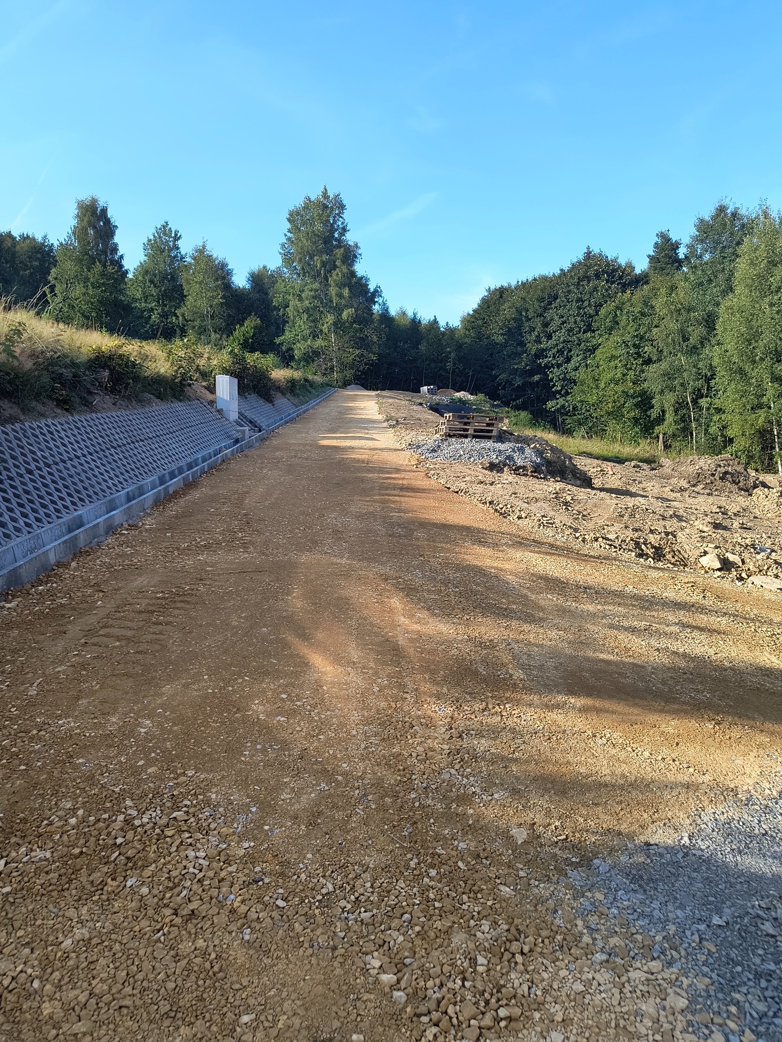 Przygotowany teren pod budowę drogi z widocznym systemem drenażowym, otoczony drzewami i błękitnym niebem.