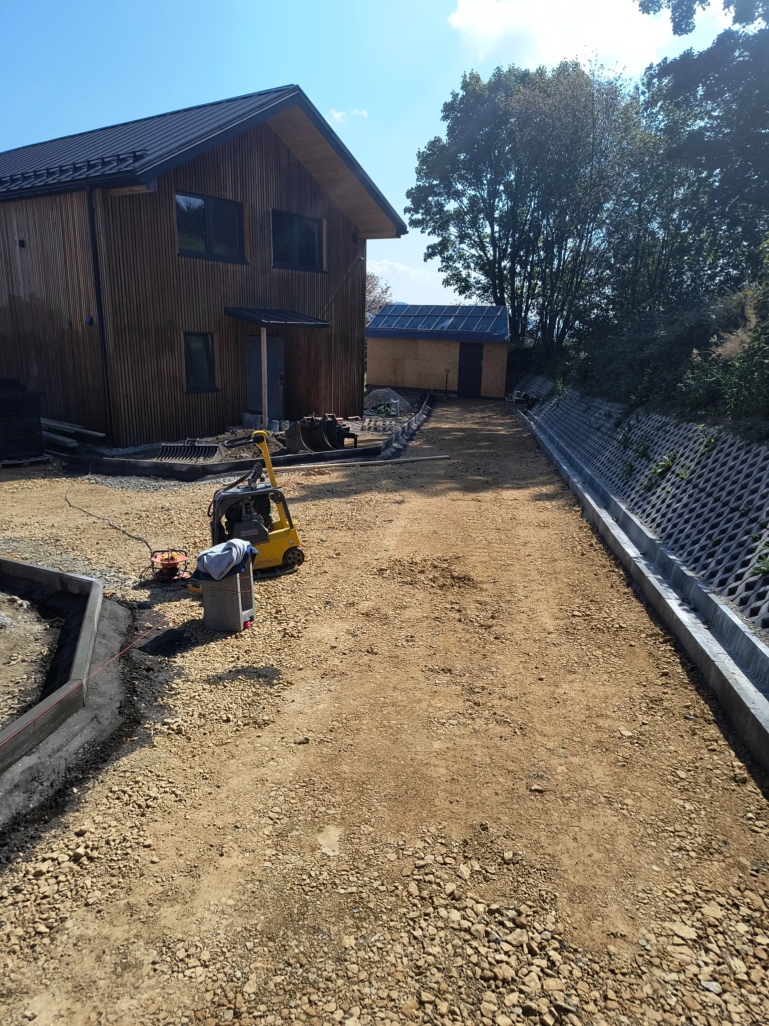 Przygotowanie podłoża pod drogę dojazdową do domu z drewnianą elewacją, widoczny zagęszczarka gruntu, krawężniki i elementy drenażu.