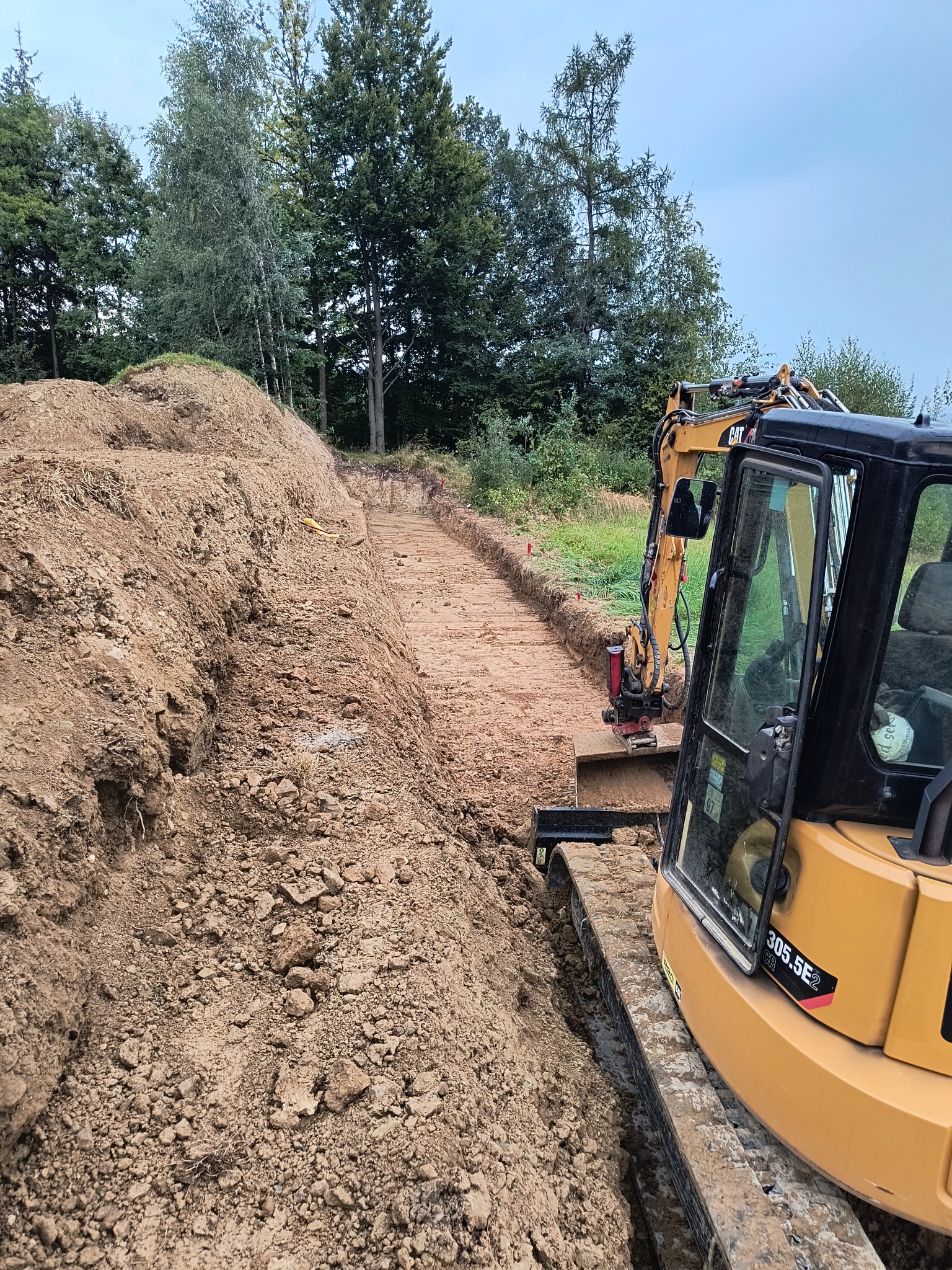 Żółta koparka gąsienicowa CAT 305.5E2 w trakcie prac ziemnych, widoczny wykop w gruncie, w tle zieleń drzew