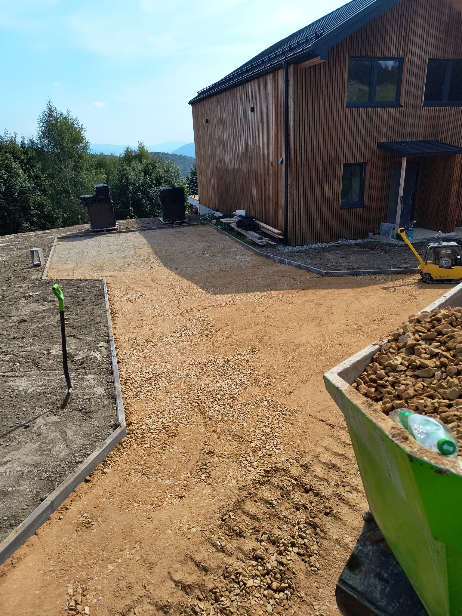 Przygotowanie podłoża z kruszywa przed nowoczesnym domem z drewnianą elewacją, widoczna ubijarka i krawężniki, w tle zalesione wzgórza.