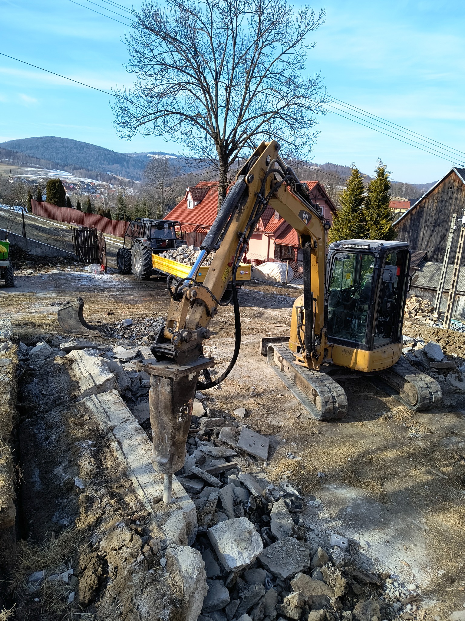 Żółty minikoparka z młotem pneumatycznym kruszy betonowy fundament, w tle traktor z przyczepą i zabudowania wiejskie.