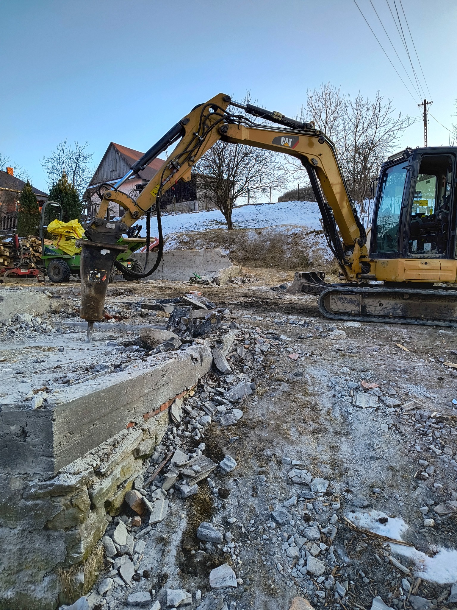 Żółty koparko-ładowarka CAT z hydraulicznym młotem pneumatycznym rozbija betonową konstrukcję; w tle widać budynki mieszkalne i resztki śniegu.
