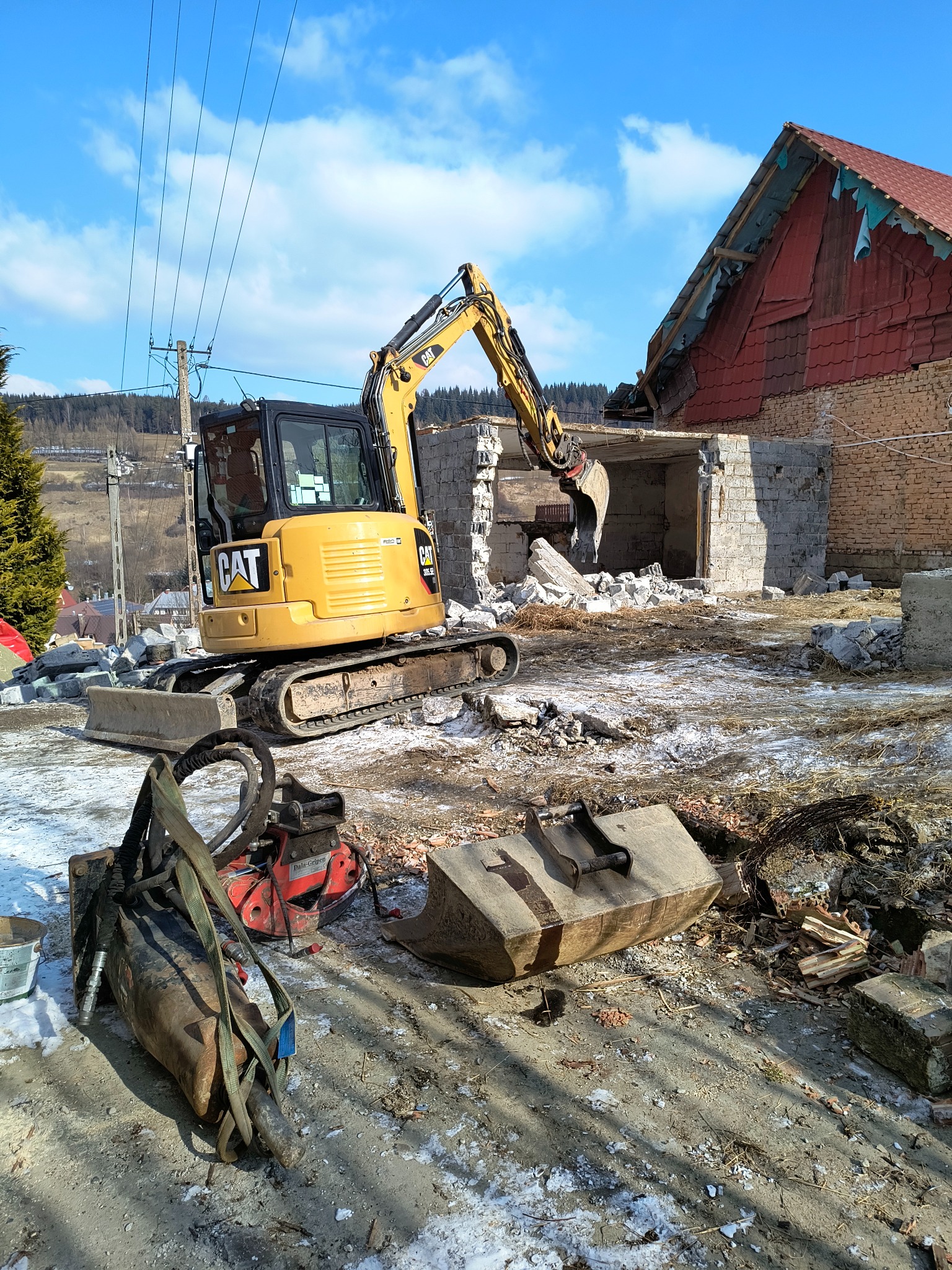 Żółta koparka CAT na placu budowy podczas rozbiórki murowanego budynku, w tle widoczny budynek z czerwoną dachówką i zalesione wzgórza pod błękitnym niebem.
