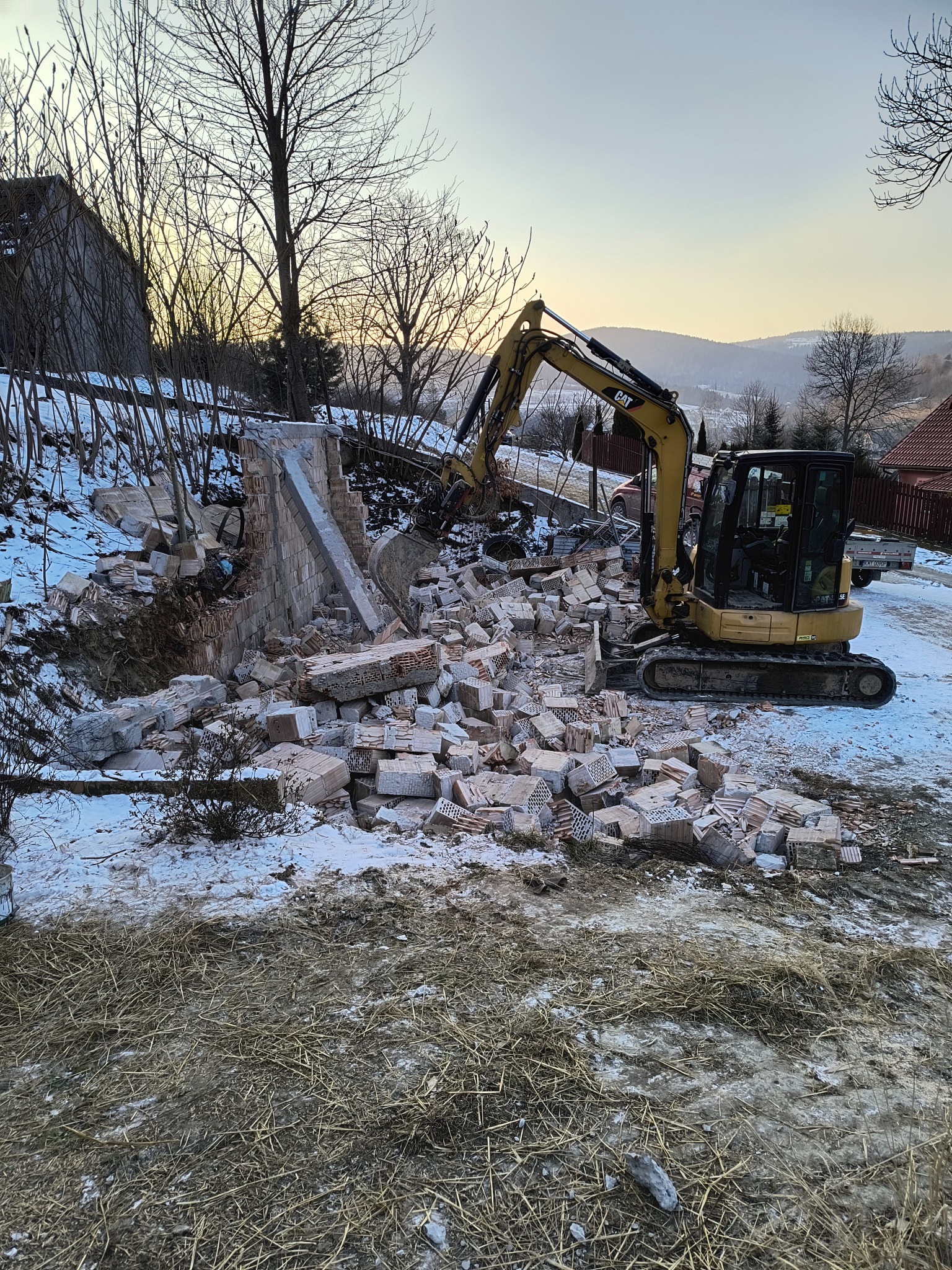 Żółta minikoparka CAT w trakcie rozbiórki murowanej konstrukcji na zaśnieżonym terenie, z widocznymi górami w tle i zachodzącym słońcem.
