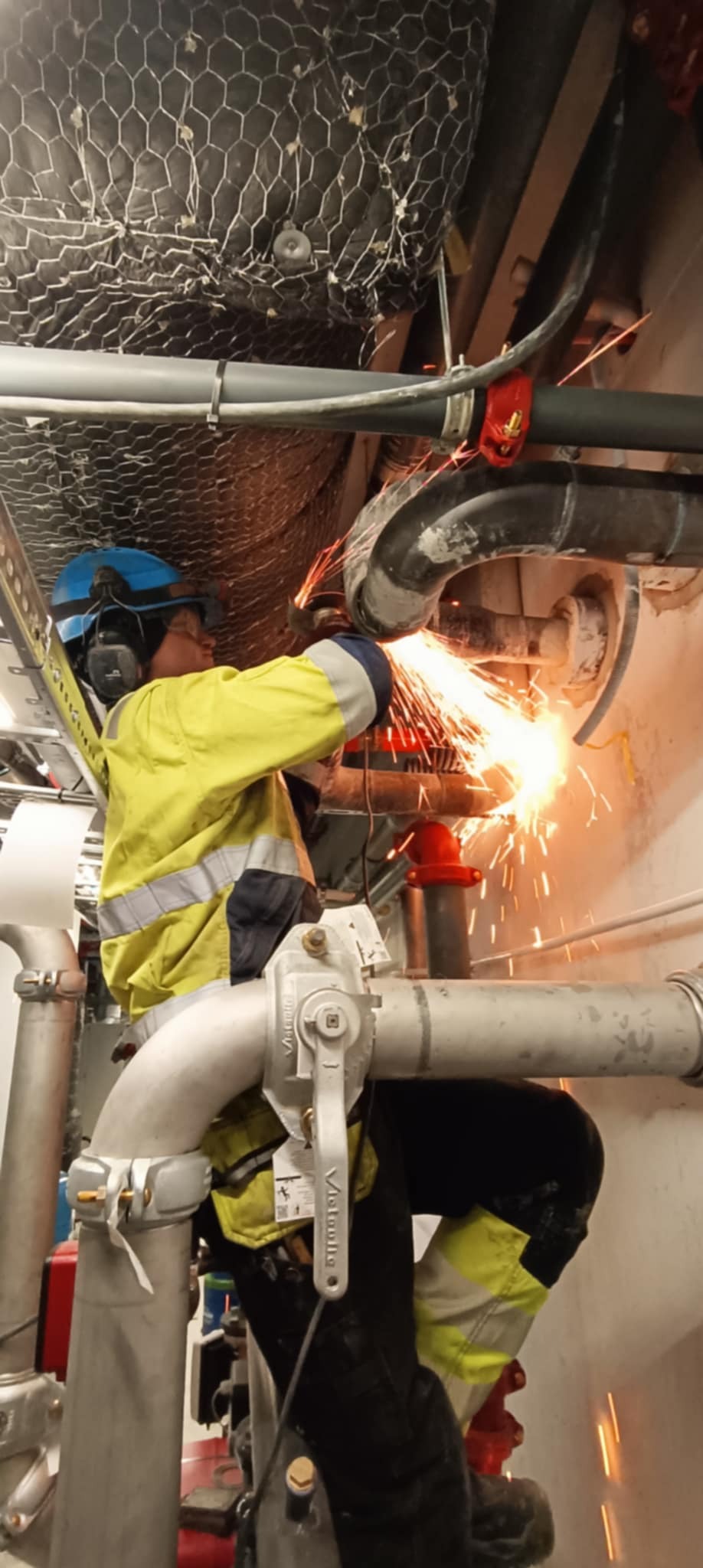 Pracownik w kasku i słuchawkach ochronnych szlifuje metalową rurę instalacji, iskry rozbryzgują się dookoła, widoczny fragment instalacji pod sufitem.