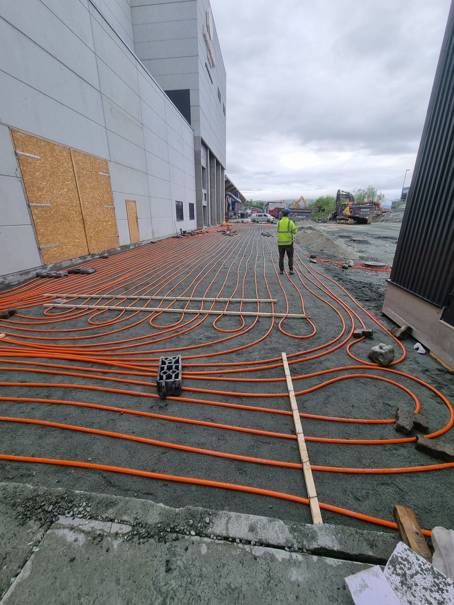 Montaż pomarańczowych rur ogrzewania podłogowego na zewnątrz budynku, pracownik w odblaskowej kamizelce nadzoruje ułożenie rur, w tle sprzęt budowlany.