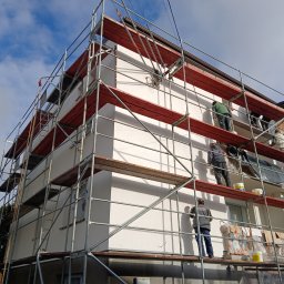 BUDOFORM - Prace na rusztowaniu podczas ocieplania ściany budynku wielorodzinnego z nową, białą elewacją. Widoczne wiadra z farbą i materiały izolacyjne.