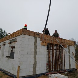 BUDOFORM - Budowa domu jednorodzinnego z bloczków, trzech robotników na stropie z szalunkiem, jeden obsługuje pompę do betonu, pochmurne niebo w tle.