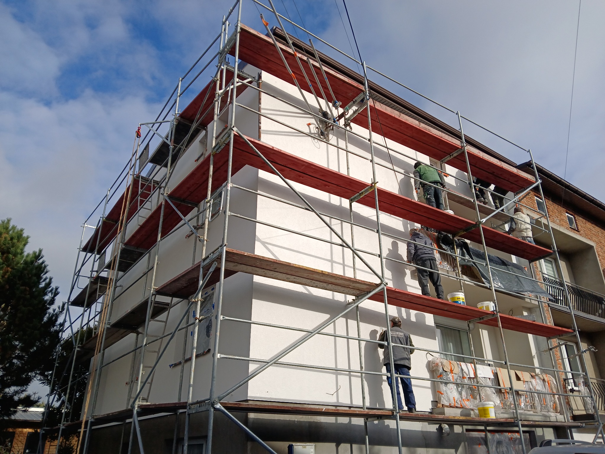 Prace na rusztowaniu podczas ocieplania ściany budynku wielorodzinnego z nową, białą elewacją. Widoczne wiadra z farbą i materiały izolacyjne.