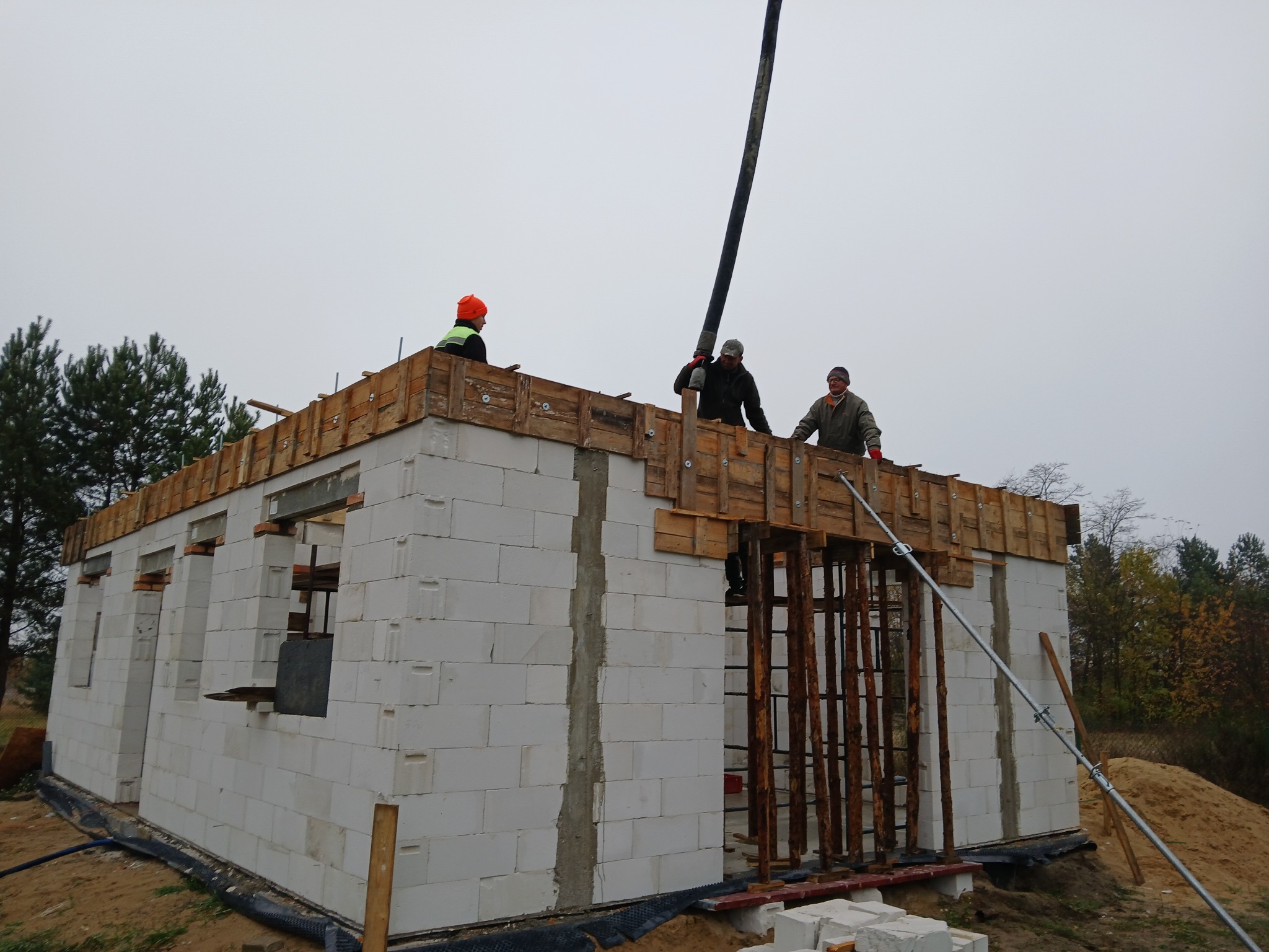 Budowa domu jednorodzinnego z bloczków, trzech robotników na stropie z szalunkiem, jeden obsługuje pompę do betonu, pochmurne niebo w tle.