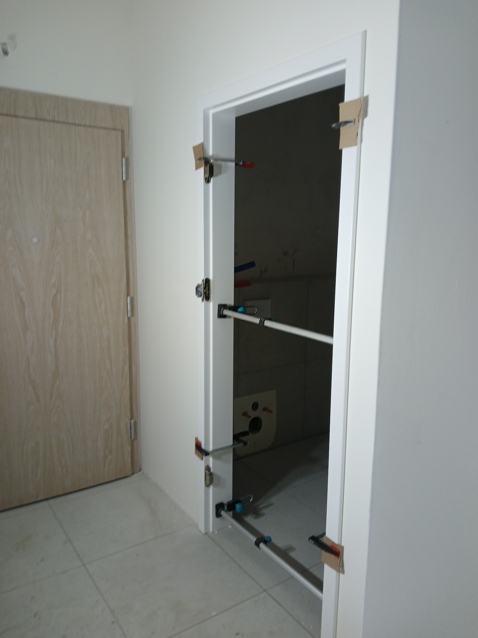 Montaż ościeżnicy drzwiowej w łazience, widoczne rozpórki teleskopowe stabilizujące ramę, zarys białej ceramiki sanitarnej w tle.