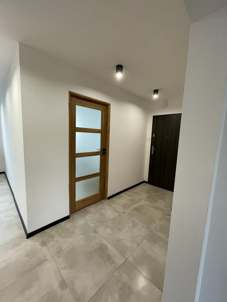 Nowoczesny przedpokój z szarą, kwadratową podłogą, drewnianą wewnętrzną i ciemną zewnętrzną stolarką drzwiową oraz minimalistycznym oświetleniem punktowym.