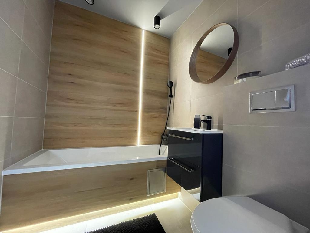 Nowoczesna łazienka z wanną obudowaną drewnopodobnymi panelami, podświetleniem LED, owalnym lustrem i czarną szafką pod umywalką.