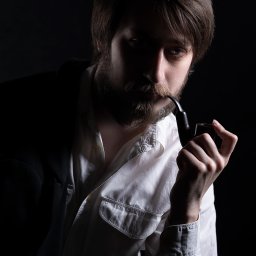 Herbata Photo - Portret mężczyzny z brodą w Toruniu, trzymającego fajkę w dłoni, w białej koszuli i marynarce na ciemnym tle.