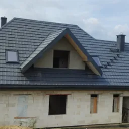 Dom w trakcie budowy z ciemnoszarym dachem z dachówki metalowej, widoczne okna dachowe i kominy na tle zalesionych wzgórz.