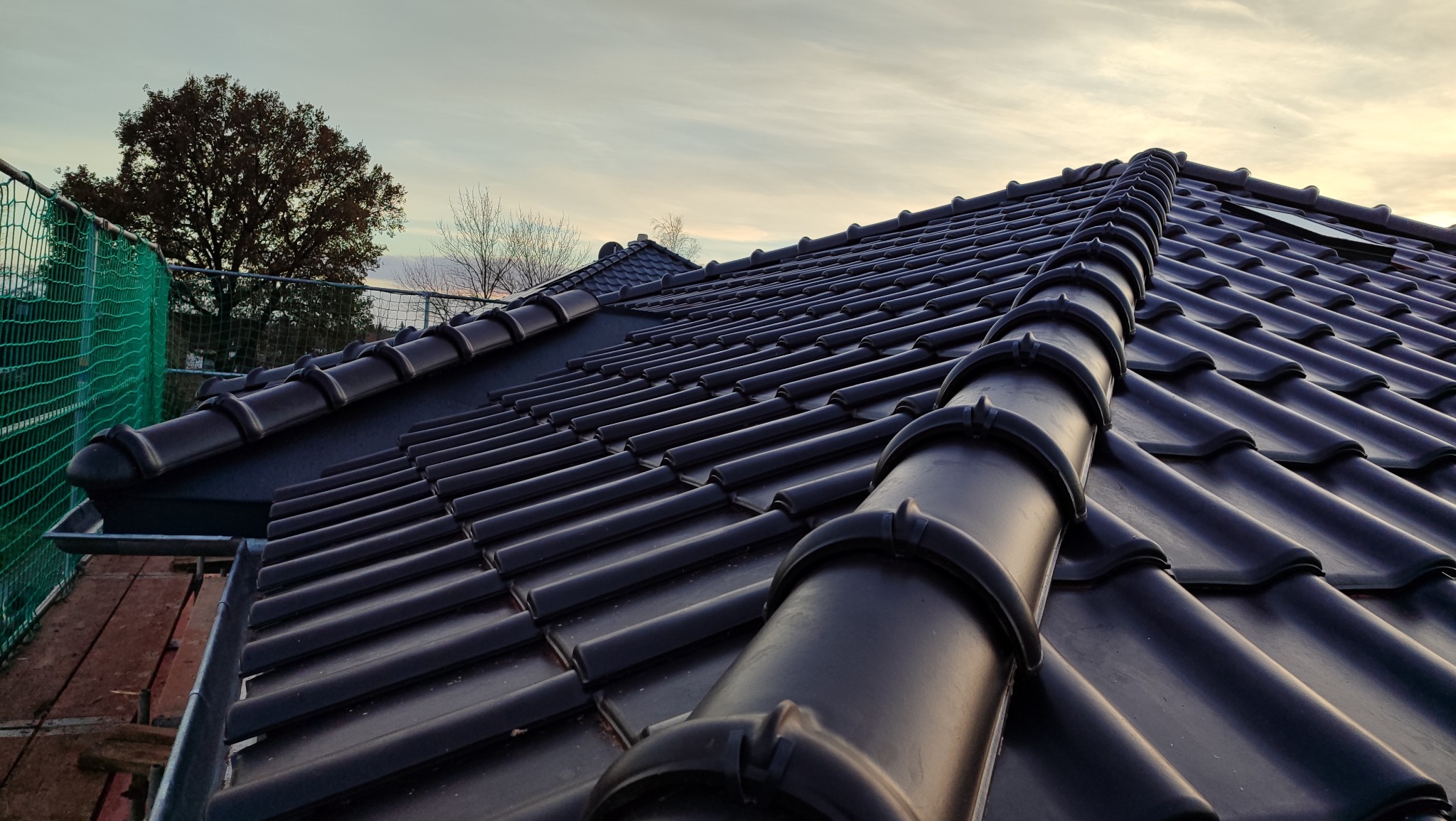 Nowy, czarny dach z ceramicznej dachówki karpiówki, widok pod kątem. W tle drzewo i siatka zabezpieczająca. Dachówka ułożona z precyzją, widoczne detale.