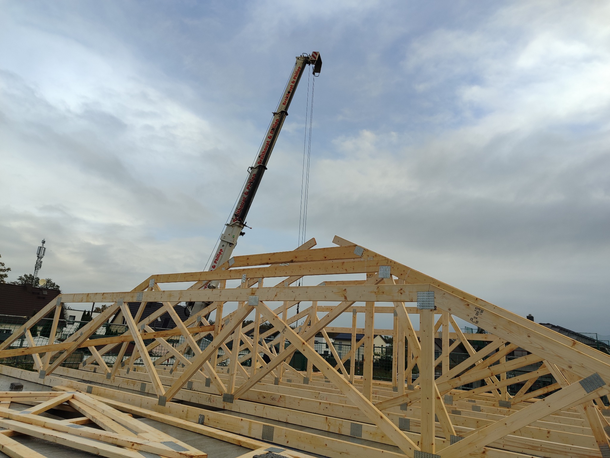 Konstrukcja drewnianej więźby dachowej w trakcie montażu z użyciem dźwigu na tle pochmurnego nieba. Widoczne elementy złączne i surowe drewno.