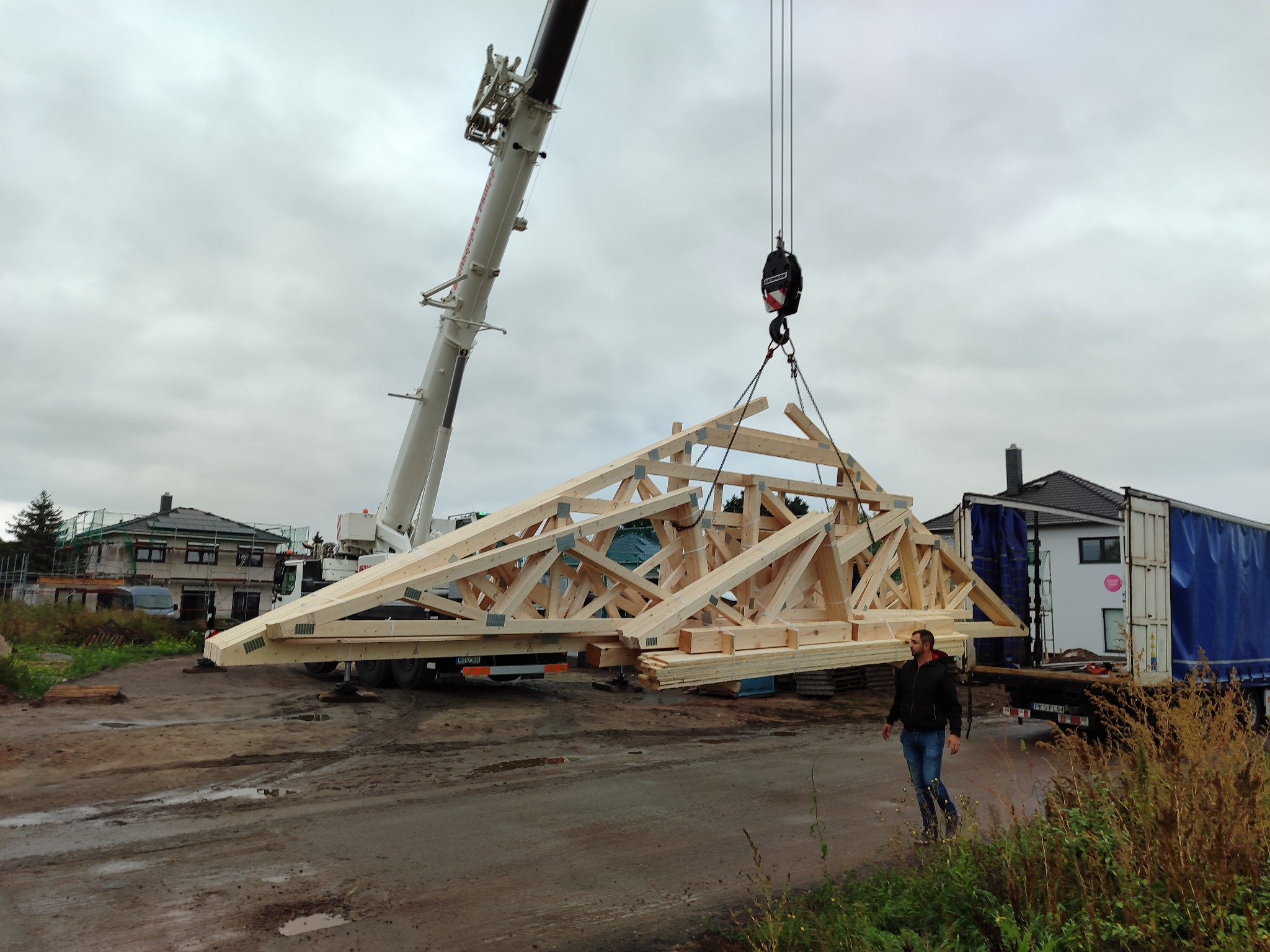 Montaż drewnianej więźby dachowej za pomocą dźwigu na placu budowy, widoczny mężczyzna w oddali. Tło stanowi częściowo wykończony dom i ciężarówka.