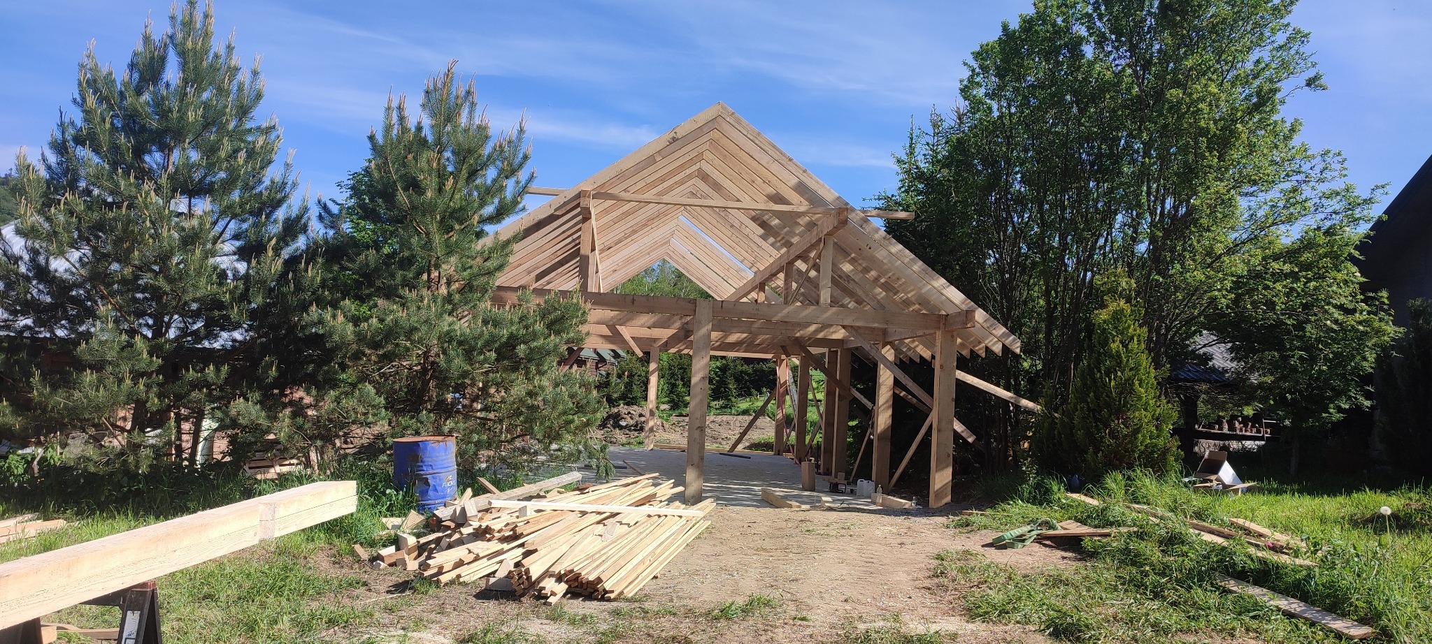 Wznoszenie drewnianej konstrukcji dachu na tle zieleni. Widoczne elementy więźby dachowej i stos desek. Budowa w toku, naturalne otoczenie.