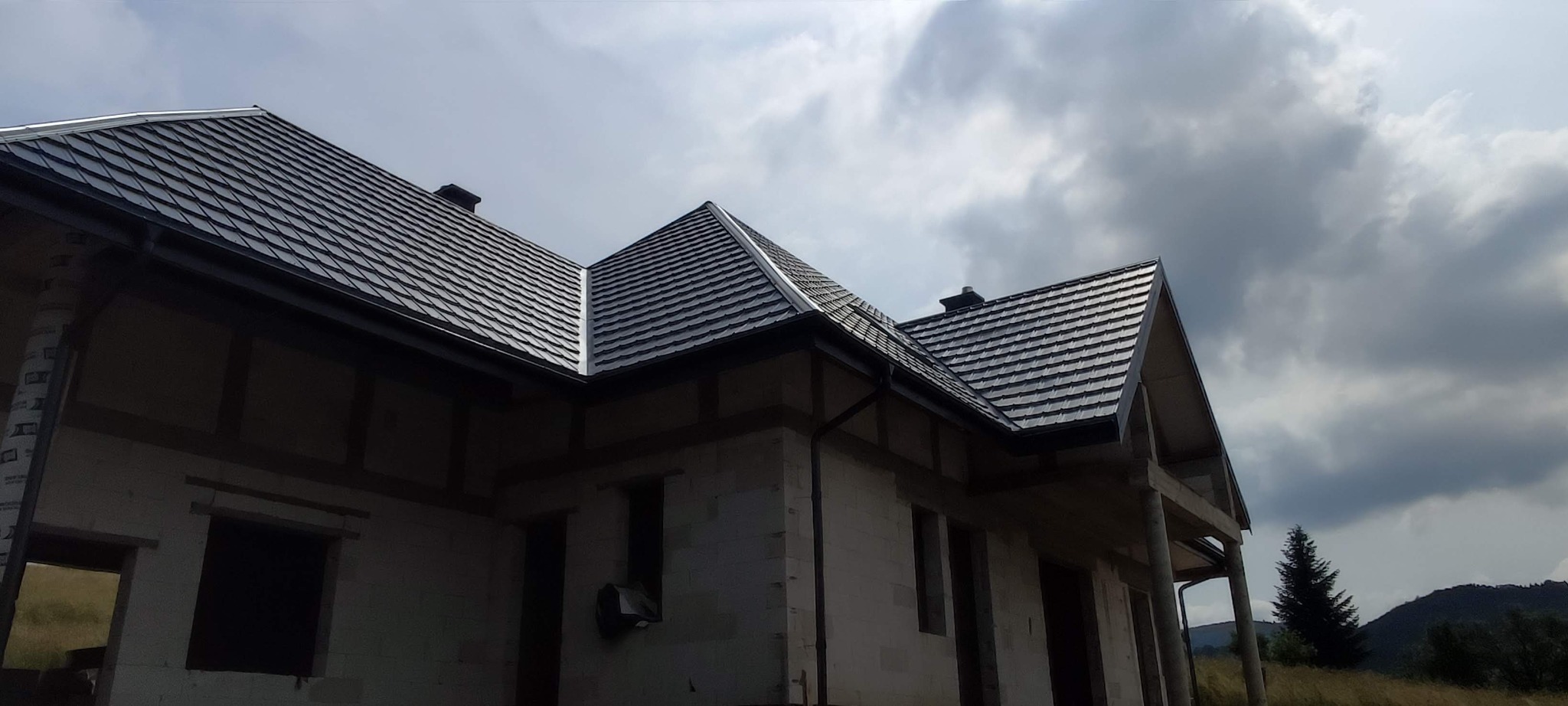 Dom w trakcie budowy z ciemnoszarą dachówką karpiówką. Widoczne obróbki blacharskie i orynnowanie. W tle pochmurne niebo i zalesione wzgórza.