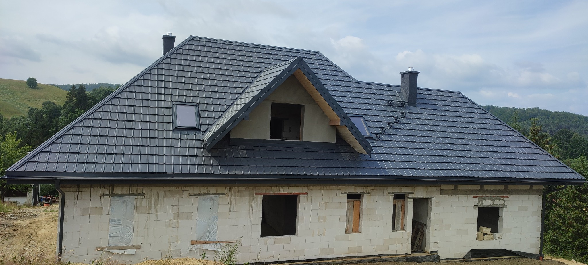 Dom w trakcie budowy z ciemnoszarym dachem z dachówki metalowej, widoczne okna dachowe i kominy na tle zalesionych wzgórz.