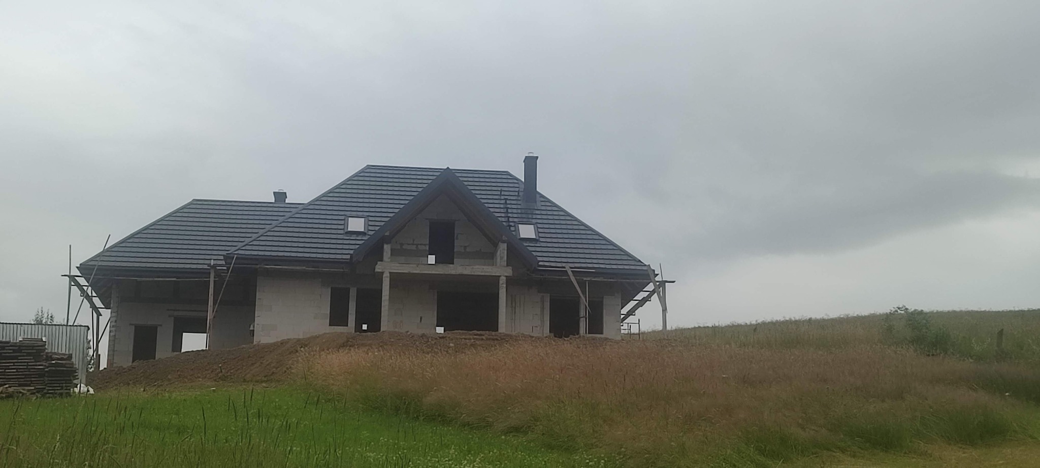 Dom w budowie z ciemną dachówką na wzgórzu, otoczony polną trawą. Widoczne rusztowania i materiały budowlane. Pochmurne niebo w tle.