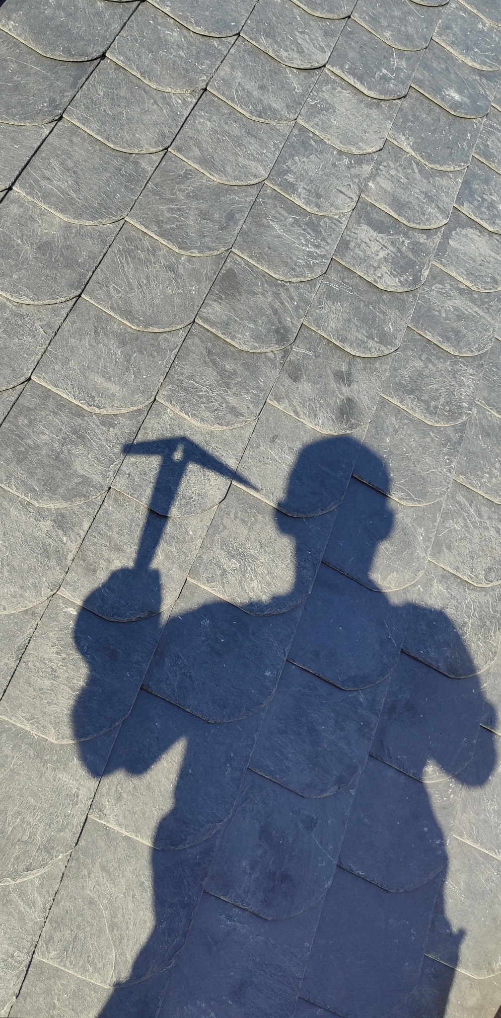 Cieniowana powierzchnia dachu pokryta łupkiem z widocznym cieniem dekarza trzymającego młotek dekarski. Nietypowe ujęcie z góry.