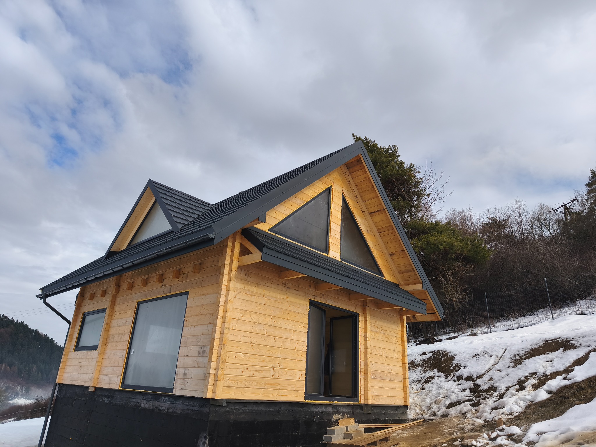 Drewniany dom z ciemnoszarym dachem z blachy na tle pochmurnego nieba. Widoczne okna i drzwi. Częściowo ośnieżony teren. Dom w stylu skandynawskim.