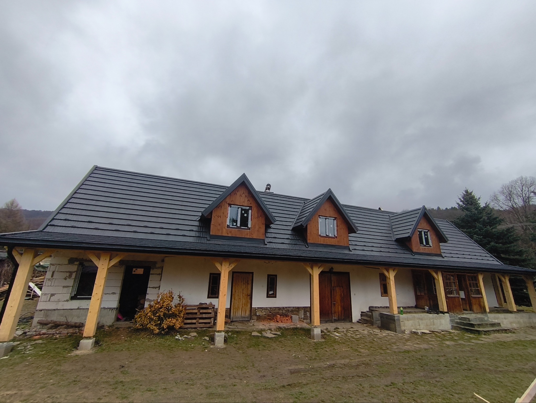Dom z ciemną dachówką ceramiczną i drewnianymi elementami wykończeniowymi. Widoczne trzy lukarny. Budynek w trakcie budowy, z elementami z pustaków.