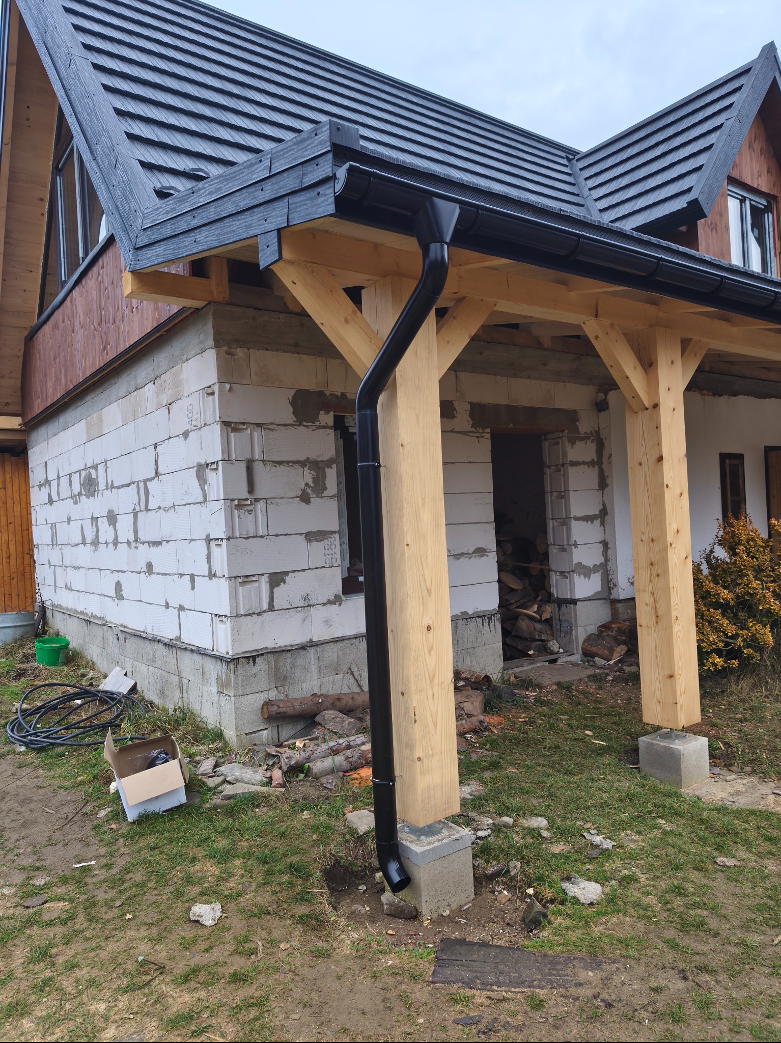 Dom w trakcie budowy z widocznym dachem krytym czarną dachówką, drewnianą konstrukcją z czarnymi rynnami i ścianami z białego bloczka. Widoczne materiały budowlane.