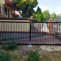 Simone Pastore - Czarna, dwuskrzydłowa brama wjazdowa z pionowymi prętami, osadzona na betonowym fundamencie, widoczna na tle domu w trakcie remontu i zadbanego ogrodu.