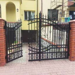 Otwarta, czarna, kuta brama wjazdowa z dekoracyjnymi elementami na tle żółtego domu z ceglanymi słupkami, widok z zewnątrz.