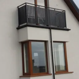 Nowoczesny balkon z czarną, pionową balustradą na tle białej elewacji domu z oknami w drewnianych ramach.
