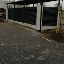 Simone Pastore - Nowoczesne, czarne ogrodzenie z poziomych paneli zadaszone, wsparte na białych słupkach, na tle szarej kostki brukowej i drzew bez liści.