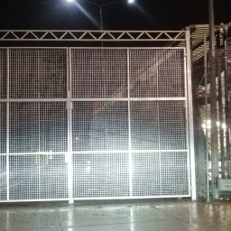 Simone Pastore - Duża, dwuskrzydłowa brama wjazdowa z metalowej siatki, widoczna nocą przy sztucznym oświetleniu, z konstrukcją nośną z profili stalowych.