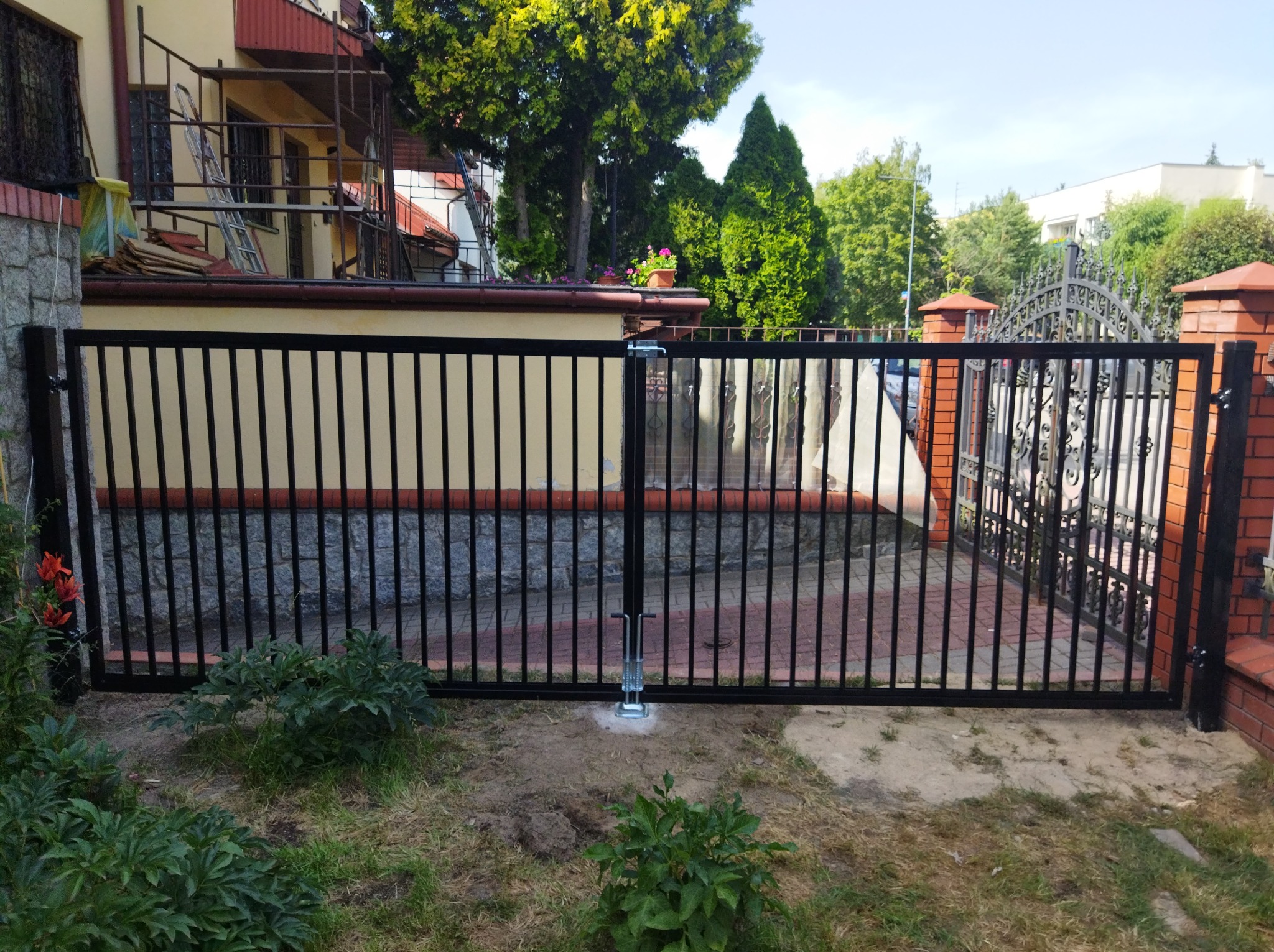 Czarna, dwuskrzydłowa brama wjazdowa z pionowymi prętami, osadzona na betonowym fundamencie, widoczna na tle domu w trakcie remontu i zadbanego ogrodu.