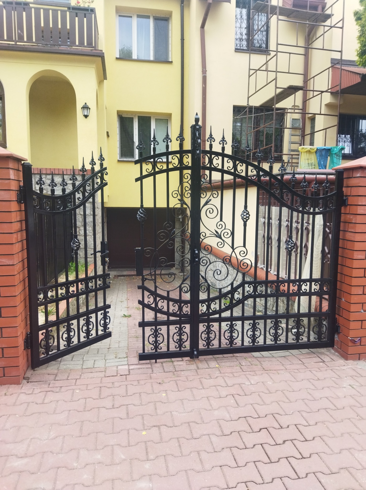 Otwarta, czarna, kuta brama wjazdowa z dekoracyjnymi elementami na tle żółtego domu z ceglanymi słupkami, widok z zewnątrz.