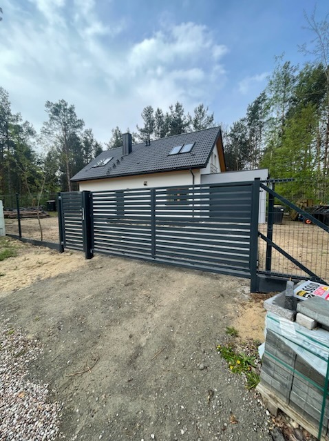 Nowoczesna, przesuwna brama wjazdowa z poziomymi panelami, częściowo otwarta, na tle domu z ciemną dachówką i lasu.