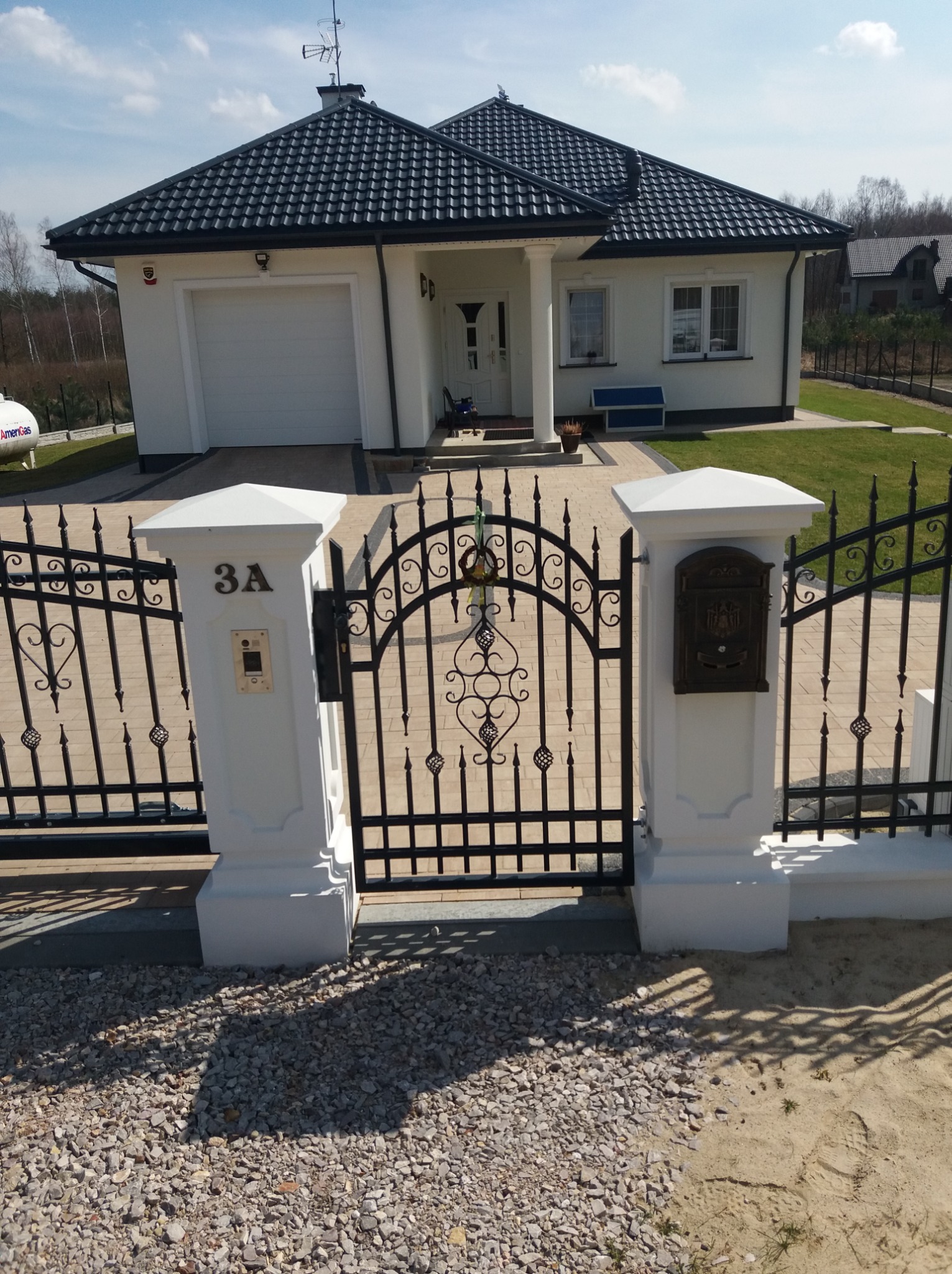 Czarna, ozdobna brama wjazdowa z numerem domu '3A' na białym słupku, skrzynka na listy po prawej stronie, w tle biały dom z czarnym dachem i zadbany trawnik.
