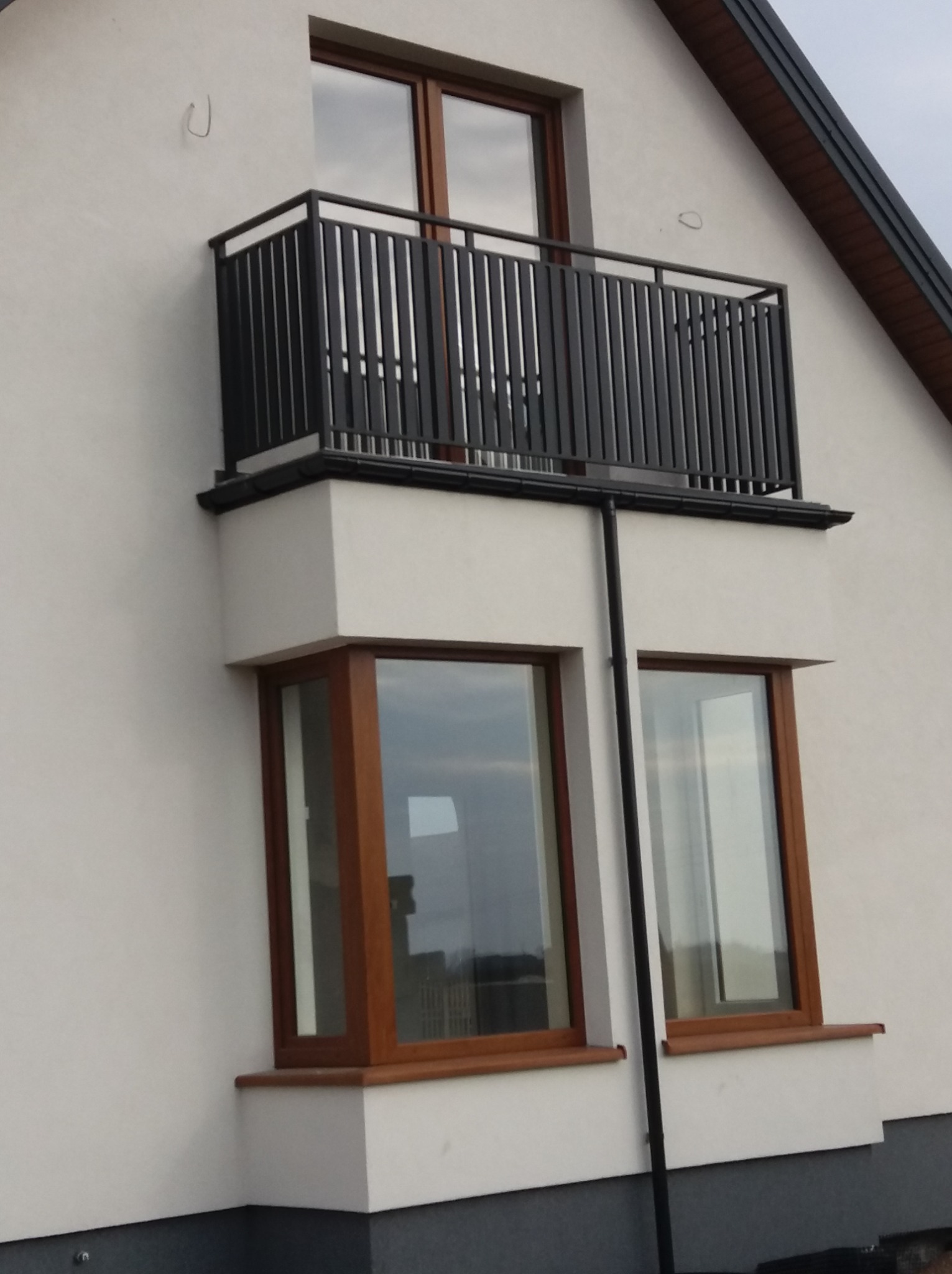 Nowoczesny balkon z czarną, pionową balustradą na tle białej elewacji domu z oknami w drewnianych ramach.