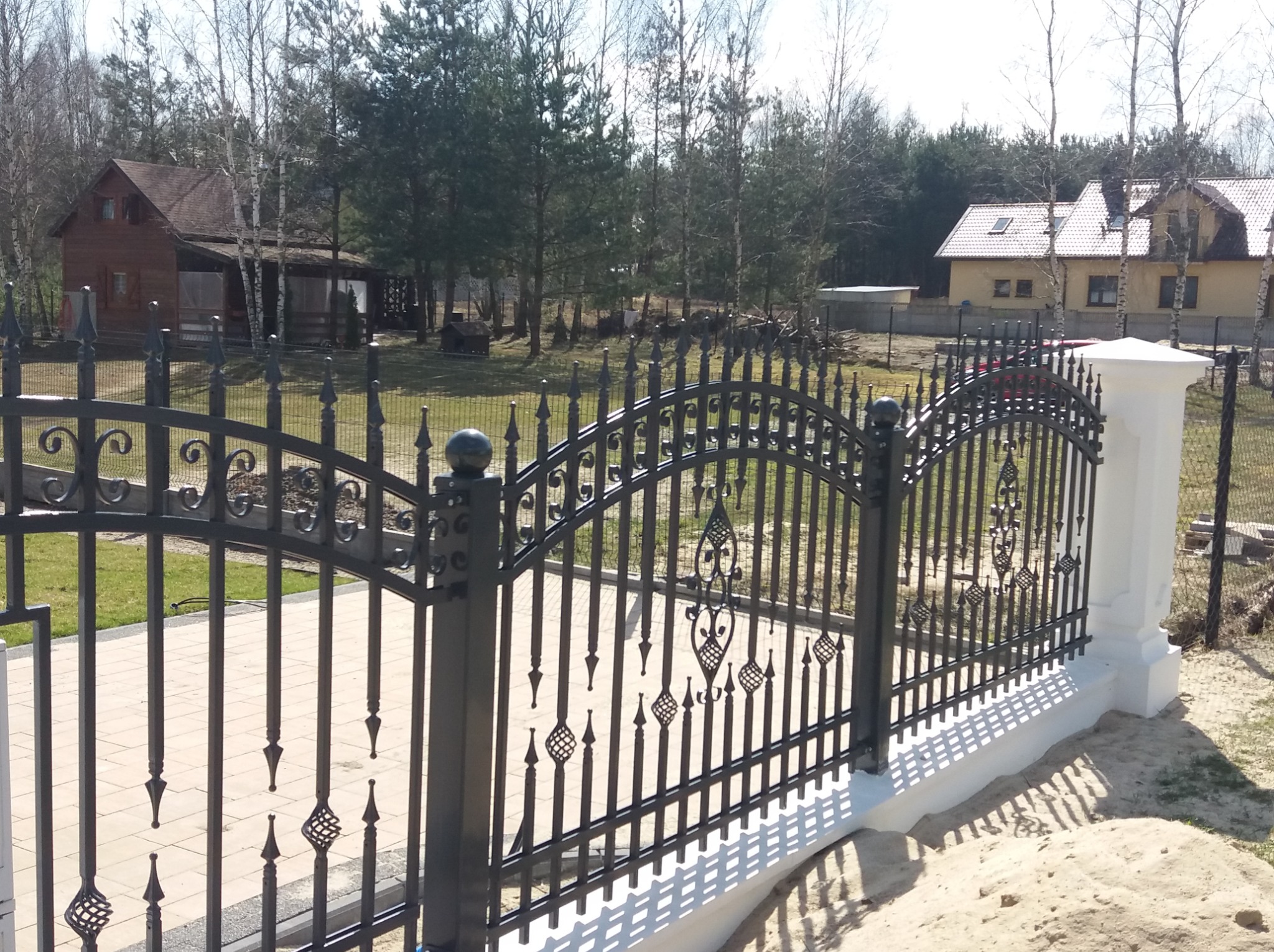 Czarny, metalowy płot z dekoracyjnymi elementami w kształcie grotów i spirali, na niskim, białym murku, z widokiem na dwa domy i drzewa w tle.