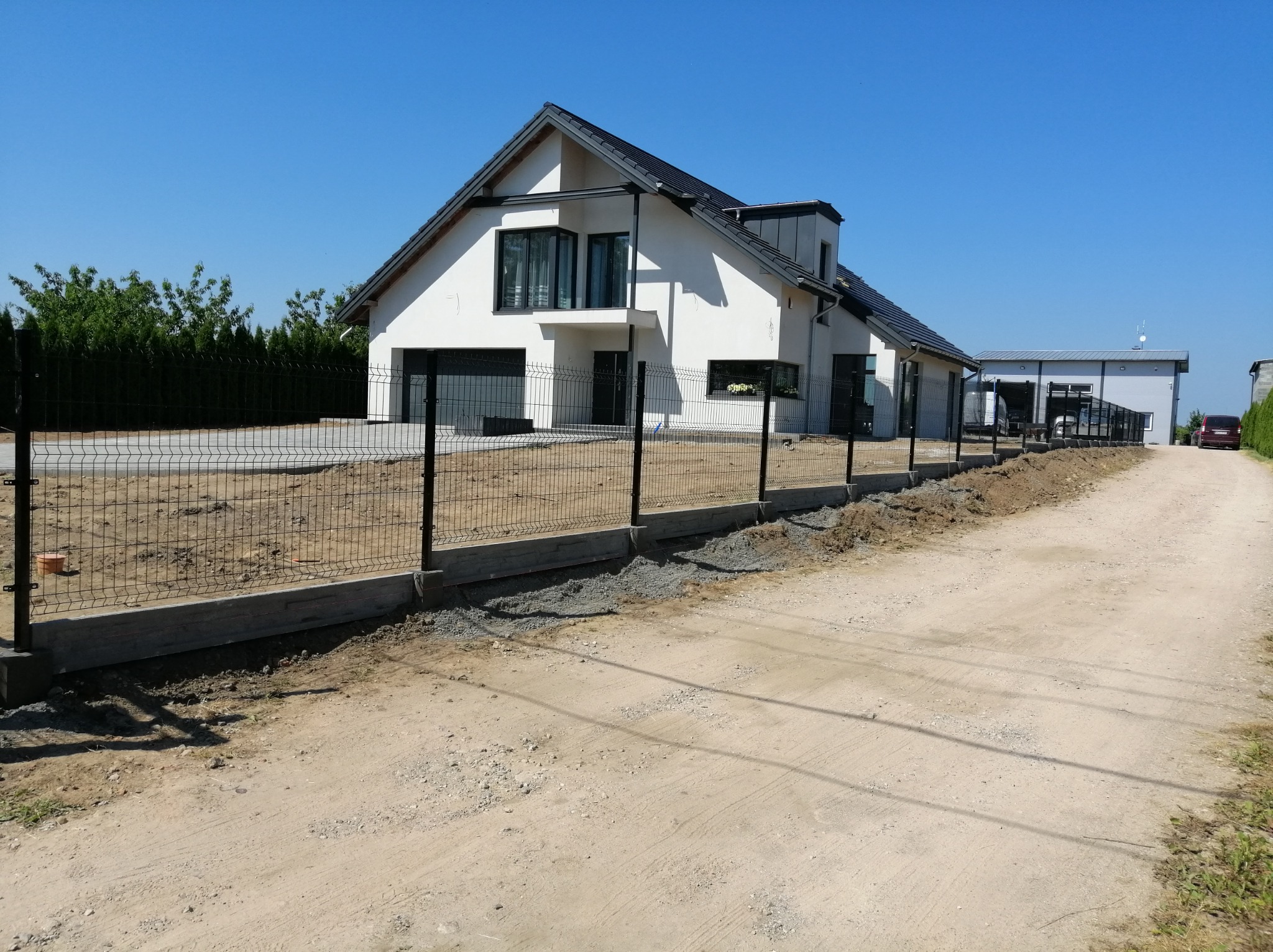 Czarny panel ogrodzeniowy z podmurówką betonową przed nowoczesnym, białym domem z garażem, widok z polnej drogi w słoneczny dzień.