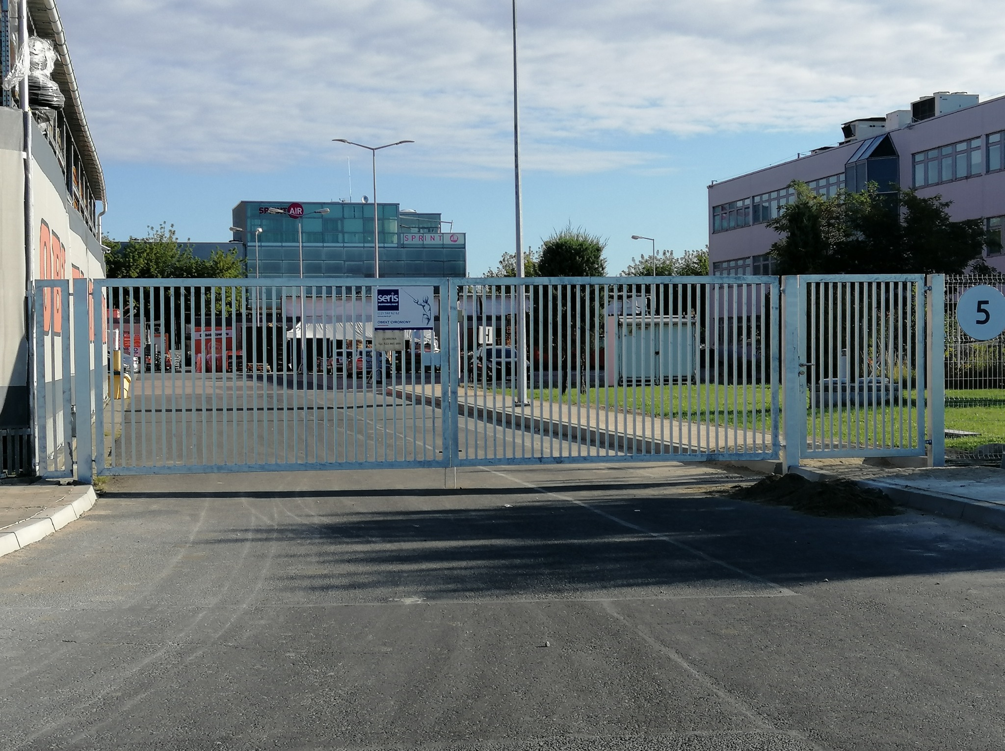 Szeroka, metalowa brama wjazdowa z pionowymi szczeblami, otwierana przesuwnie, z tabliczką informacyjną firmy ochroniarskiej, na tle budynków biurowych w słoneczny dzień.