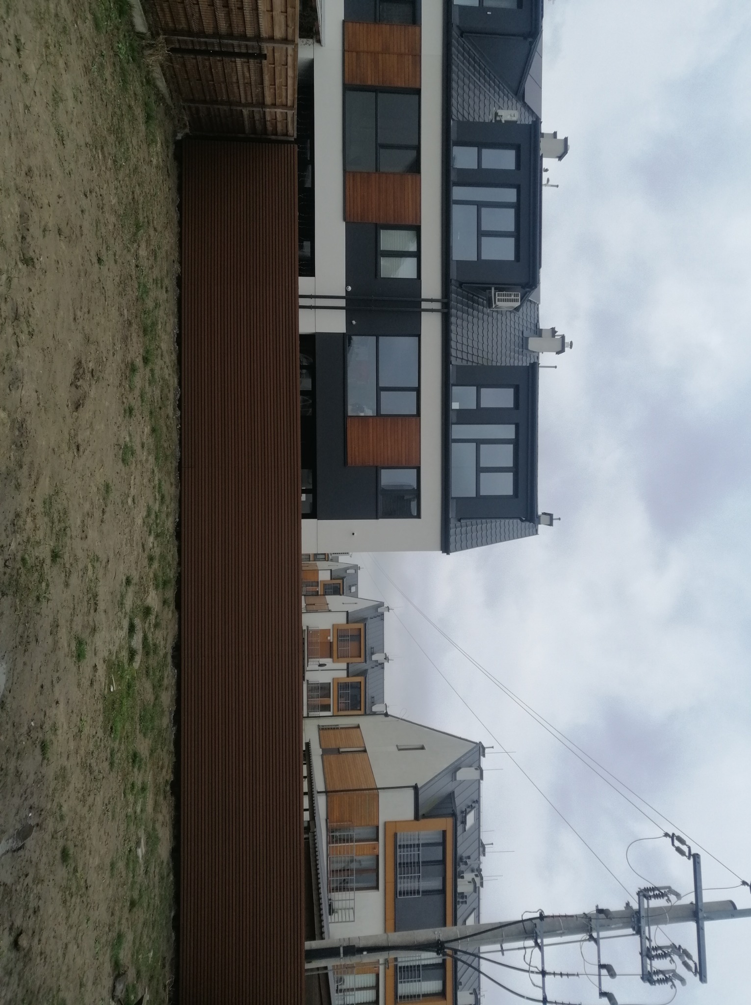 Nowoczesne budynki wielorodzinne z panelowym ogrodzeniem w kolorze ciemnego drewna, widoczne słupy energetyczne i niebo z chmurami.