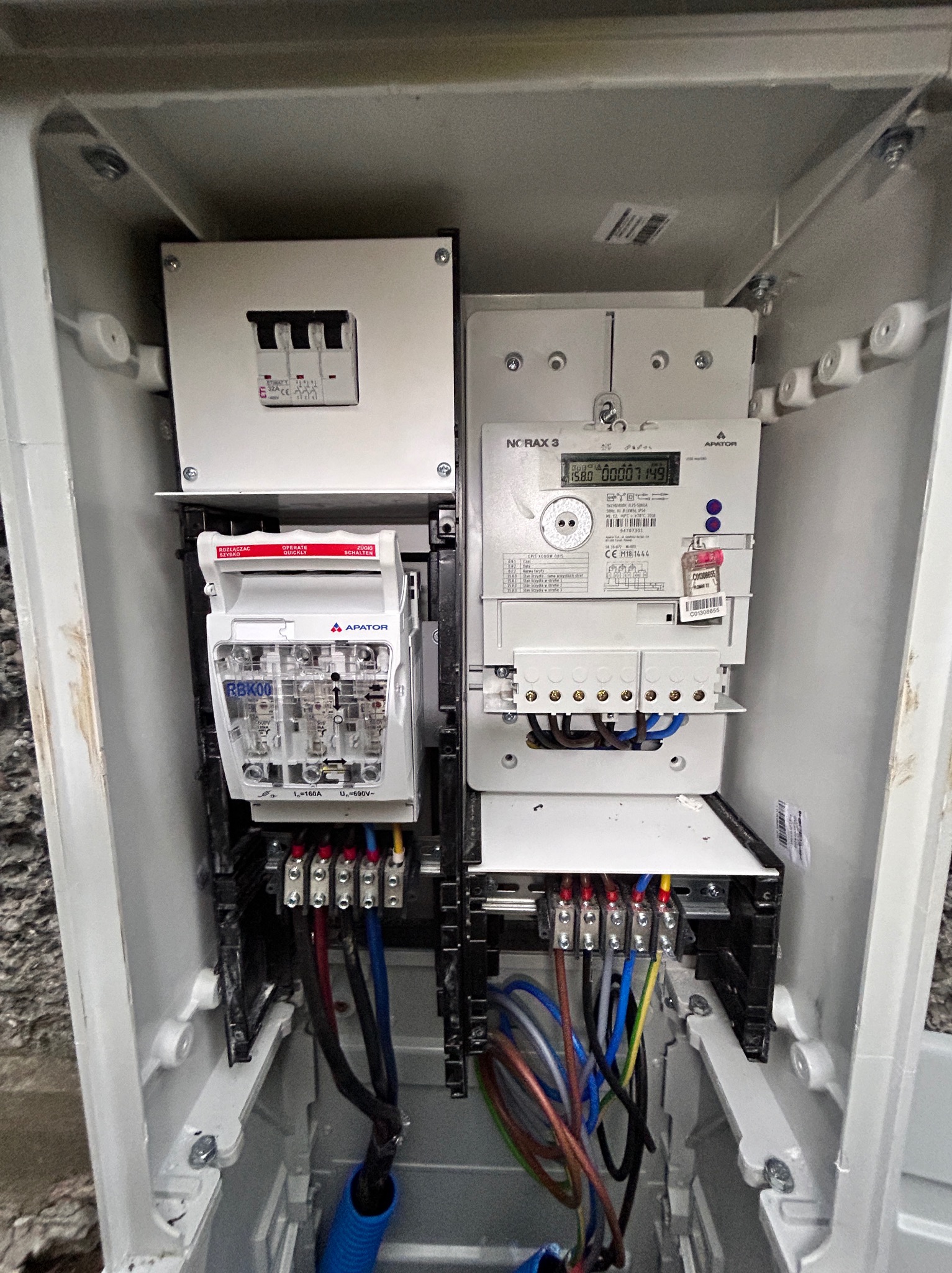 Wnętrze skrzynki elektrycznej z zamontowanym licznikiem energii, rozłącznikiem bezpiecznikowym RBK00 marki Apator i zabezpieczeniami nadprądowymi, widoczne podłączenia przewodów zasilających.