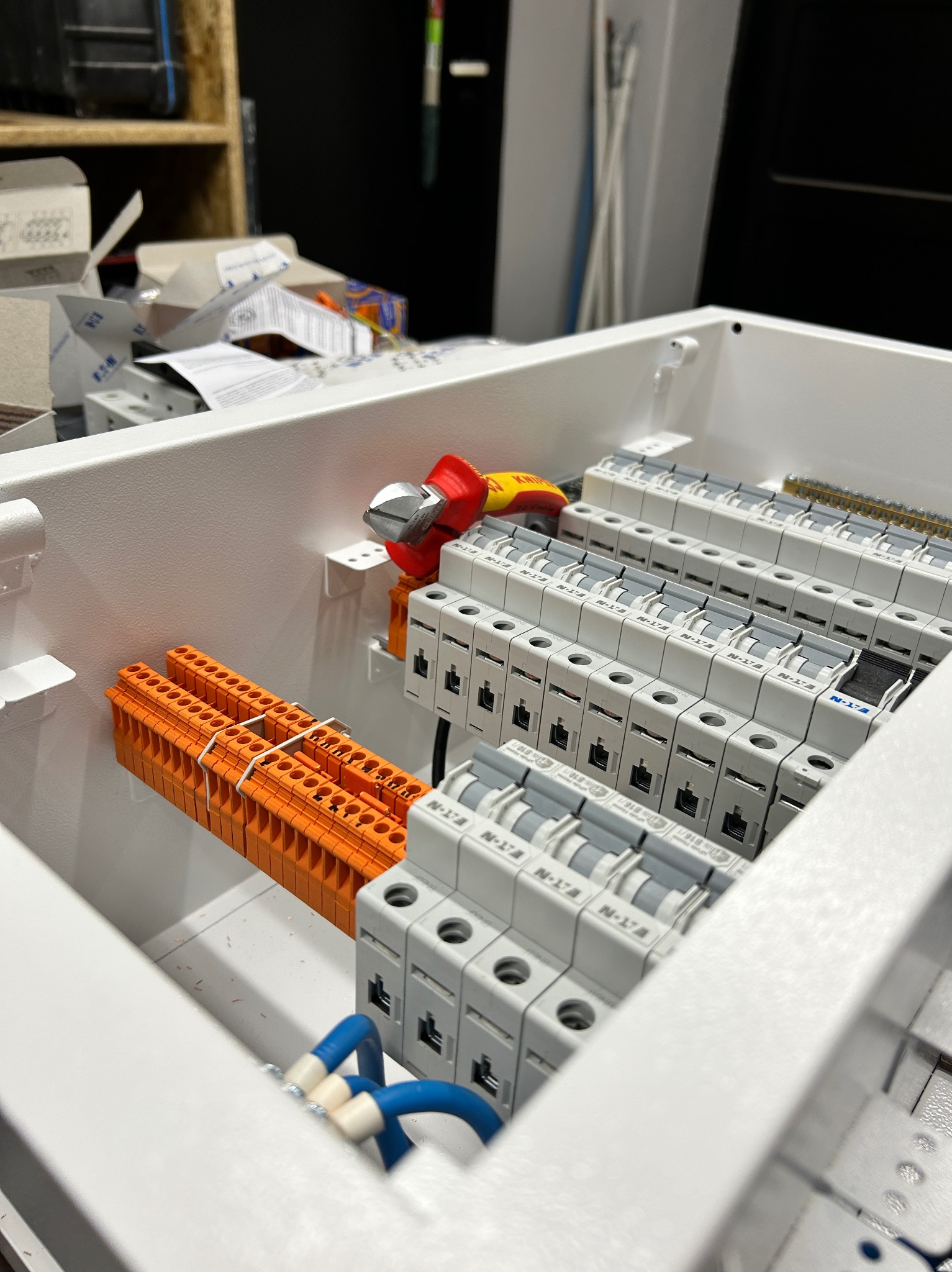 Wnętrze skrzynki elektrycznej z zamontowanymi bezpiecznikami, listwami zaciskowymi i przewodami; widoczne narzędzia do cięcia przewodów.